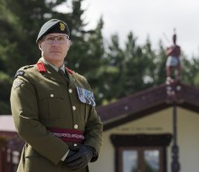 A male soldier wears army green ceremonial uniform, medals on his left breast, brown gloves, red lapel pins and a green beret.  He stands proudly in front of a marae and pine trees at the rear.