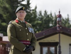 A male soldier wears army green ceremonial uniform, medals on his left breast, brown gloves, red lapel pins and a green beret.  He stands proudly in front of a marae and pine trees at the rear.