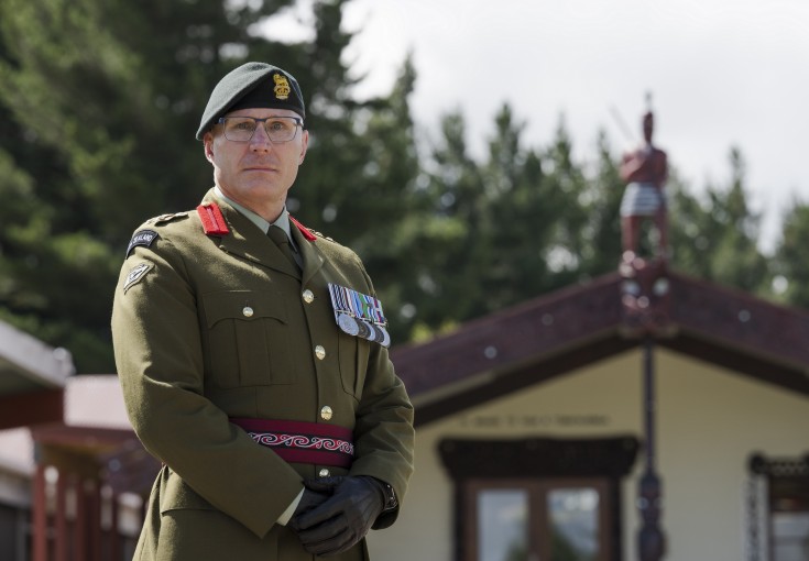 A male soldier wears army green ceremonial uniform, medals on his left breast, brown gloves, red lapel pins and a green beret.  He stands proudly in front of a marae and pine trees at the rear.