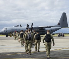 A group of soldiers walk toward a large grey military aircraft, they walk in two files, black backpacks on as military staff stand alongside with high vis vests.