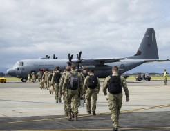 A group of soldiers walk toward a large grey military aircraft, they walk in two files, black backpacks on as military staff stand alongside with high vis vests.