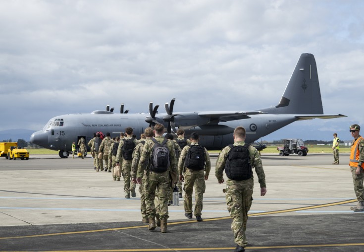 A group of soldiers walk toward a large grey military aircraft, they walk in two files, black backpacks on as military staff stand alongside with high vis vests.