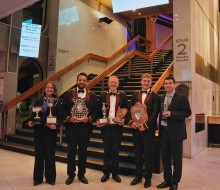 Five people dressed smartly in suits stand in front of stairs holding various trophies and shields.