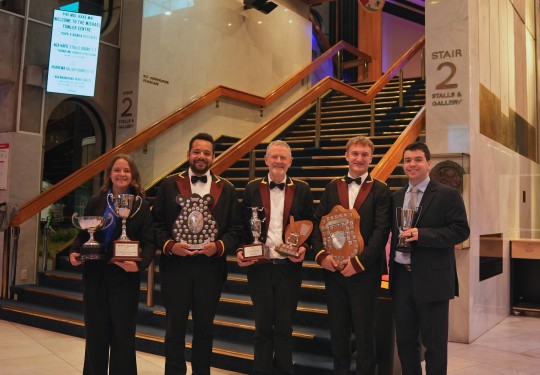 Five people dressed smartly in suits stand in front of stairs holding various trophies and shields.