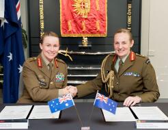 Two women Chiefs of Army for New Zealand and Australia sit at a table in front of two documents and their corresponding countries flag, they are both smiling and shaking hands. 