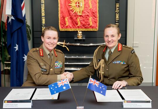 Two women Chiefs of Army for New Zealand and Australia sit at a table in front of two documents and their corresponding countries flag, they are both smiling and shaking hands. 