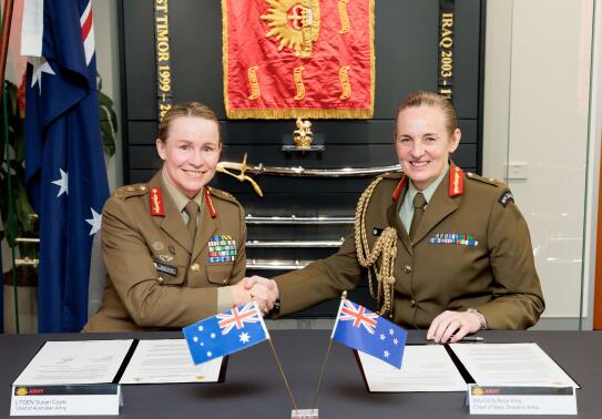 Two women Chiefs of Army for New Zealand and Australia sit at a table in front of two documents and their corresponding countries flag, they are both smiling and shaking hands. 
