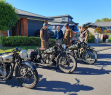 Four men stand being three vintage motorcycles on a tidy suburban street on a sunny morning.