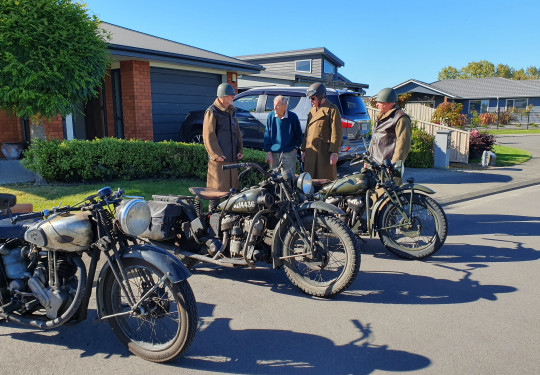 Four men stand being three vintage motorcycles on a tidy suburban street on a sunny morning.