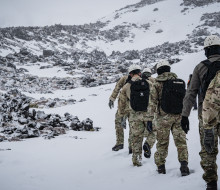Personnel walk through the snow, in uniform, in a line as part of five-day cold weather survival course on Mt Ruapehu. In the background you can see rock formation that have a dusting of snow and snow around them. 