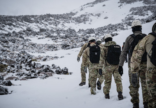 Personnel walk through the snow, in uniform, in a line as part of five-day cold weather survival course on Mt Ruapehu. In the background you can see rock formation that have a dusting of snow and snow around them. 