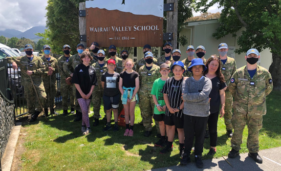 The officer cadets with some of the students and staff from Wairau Valley School. The uniformed personnel have masks on and it is a sunny day. 