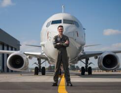 A man stands in dark green overalls with arms cross in front of the nose of a large white aircraft with a single jet engine on each wing.