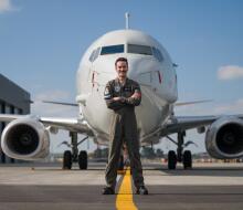 A man stands in dark green overalls with arms cross in front of the nose of a large white aircraft with a single jet engine on each wing.