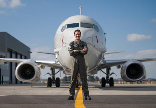 A man stands in dark green overalls with arms cross in front of the nose of a large white aircraft with a single jet engine on each wing.
