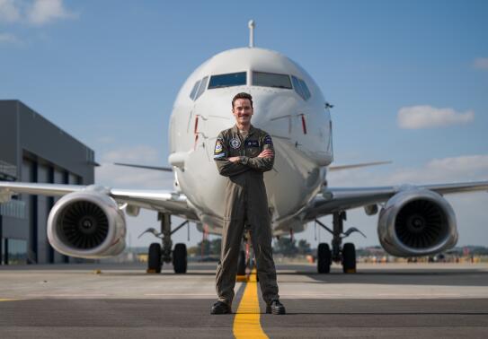 A man stands in dark green overalls with arms cross in front of the nose of a large white aircraft with a single jet engine on each wing.