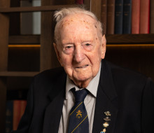A man wearing a suit and medals looks at the camera as he stands in front of a bookshelf.