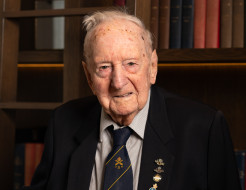 A man wearing a suit and medals looks at the camera as he stands in front of a bookshelf.