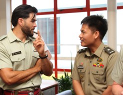 Two army officers wearing green shirts with ribbons and name badges engage in conversation standing near a window.  The taller of the two wears a watch and gestures as he talks to the other officer.