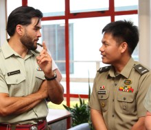 Two army officers wearing green shirts with ribbons and name badges engage in conversation standing near a window.  The taller of the two wears a watch and gestures as he talks to the other officer.