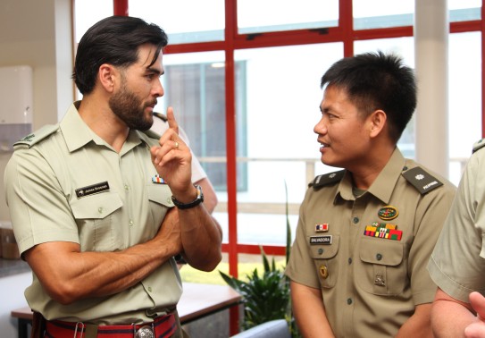 Two army officers wearing green shirts with ribbons and name badges engage in conversation standing near a window.  The taller of the two wears a watch and gestures as he talks to the other officer.