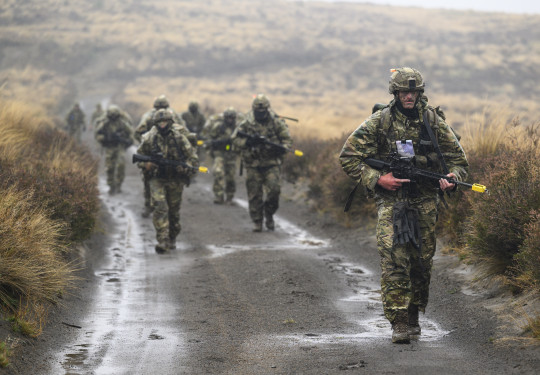 Soldiers in full uniform with weapons walk towards the camera down a wet gravel road with tussocks covering the hills in the background. 