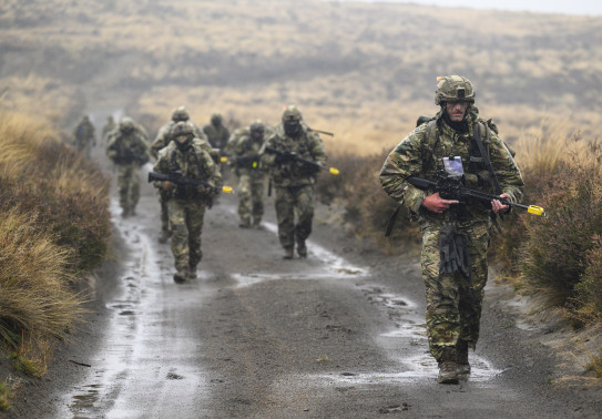 Soldiers in full uniform with weapons walk towards the camera down a wet gravel road with tussocks covering the hills in the background. 