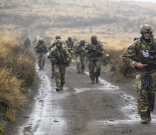 Soldiers in full uniform with weapons walk towards the camera down a wet gravel road with tussocks covering the hills in the background.