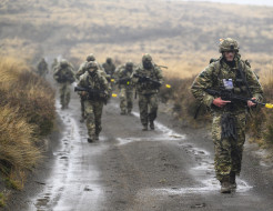 Soldiers in full uniform with weapons walk towards the camera down a wet gravel road with tussocks covering the hills in the background. 