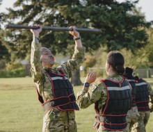 Two soldiers partner up completing a physical challenge, one soldier with a full face tattoo (Mataora) lifts a steel bar overhead, while his female counterpart raises her arms to aid if needed.  There is another two army personnel in the background who ar