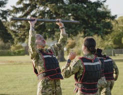 Two soldiers partner up completing a physical challenge, one soldier with a full face tattoo (Mataora) lifts a steel bar overhead, while his female counterpart raises her arms to aid if needed.  There is another two army personnel in the background who ar
