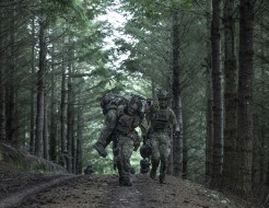 Two soldiers run through the forest canopy whilst carrying another on their soldier. They are wearing camouflage uniform, their faces are obscured 