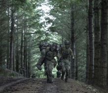 Two soldiers run through the forest canopy whilst carrying another on their soldier. They are wearing camouflage uniform, their faces are obscured 