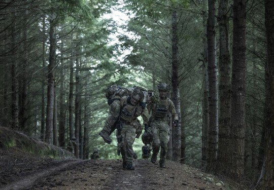 Two soldiers run through the forest canopy whilst carrying another on their soldier. They are wearing camouflage uniform, their faces are obscured 