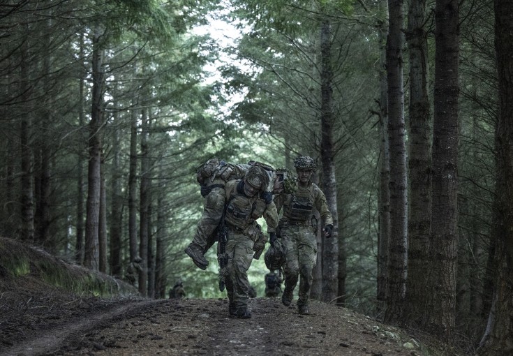 Two soldiers run through the forest canopy whilst carrying another on their soldier. They are wearing camouflage uniform, their faces are obscured 