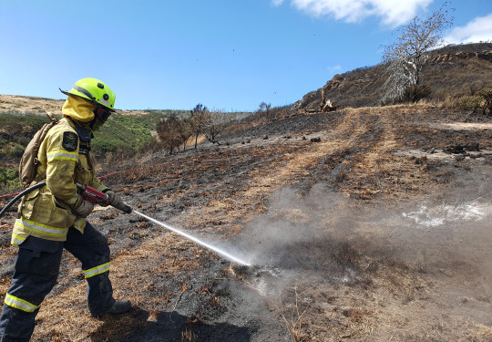 A firefighter in full PPE sprays the side of a hill, still smouldering from a fire.