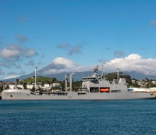 A large Naval vessel sits alongside the New Plymouth port area, it is a sunny day with blue skies and the city of New Plymouth can be seen in the background along with Mt Taranaki. The top of the mountain is obscured by cloud.