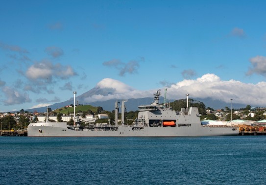A large Naval vessel sits alongside the New Plymouth port area, it is a sunny day with blue skies and the city of New Plymouth can be seen in the background along with Mt Taranaki. The top of the mountain is obscured by cloud.