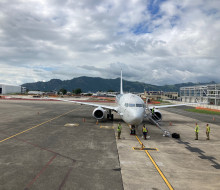 A front-on view of the RNZAF P-8A Poseidon on the tarmac in Fiji under broken clouds. In the background is the Fiji Airways hangar and a hills covered in forest.