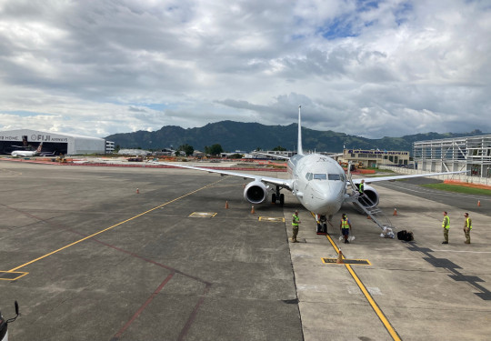 A front-on view of the RNZAF P-8A Poseidon on the tarmac in Fiji under broken clouds. In the background is the Fiji Airways hangar and a hills covered in forest.