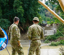 Three soldiers stand on the bank of a river where a bridge once stood. On the other side of the river, a small part of the bridge remains but the rest is gone. Debris from the high flood waters is caught in trees and on the sides of the river.