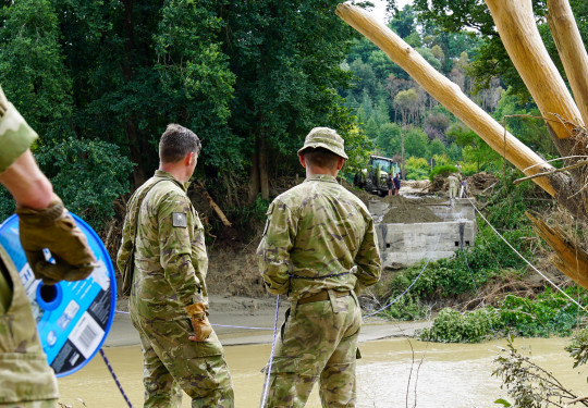 Three soldiers stand on the bank of a river where a bridge once stood. On the other side of the river, a small part of the bridge remains but the rest is gone. Debris from the high flood waters is caught in trees and on the sides of the river.