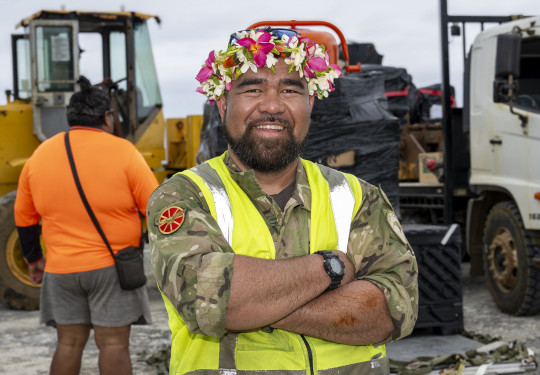 An aviator stands, arms crossed and smiling at the camera. He wears a Cook Island ei katu, a garland of flowers worn on his head, a high vis jacket and RNZAF uniform,