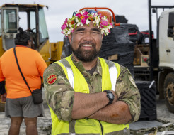 An aviator stands, arms crossed and smiling at the camera. He wears a Cook Island ei katu, a garland of flowers worn on his head, a high vis jacket and RNZAF uniform,