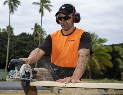 A soldier in high vis orange saws a piece of wood, The soldier is wearing a black peaked cap with an NZDF Kiwi logo on it, ear muffs and eye protection. It's a cloudy day. 