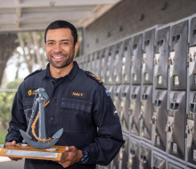Man in Navy uniform smiles as he holds a trophy that resembles an anchor.
