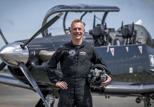 A male wearing a black flying suit holds a flying helmet in one hand whilst the other hand rests on his hip.  He grins at the camera and stands in front of a small black aircraft.