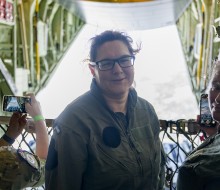 A brown haired woman with glasses, wearing a green military flying suit, looks into the camera whilst standing on board a large military aircraft.  Beside her is a military female holding a phone taking a photo of the scene behind - the open door of the a