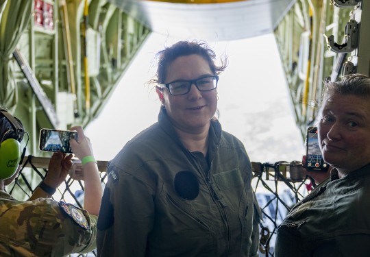 A brown haired woman with glasses, wearing a green military flying suit, looks into the camera whilst standing on board a large military aircraft.  Beside her is a military female holding a phone taking a photo of the scene behind - the open door of the a