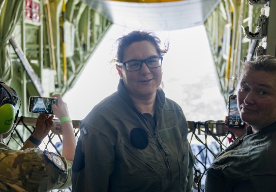 A brown haired woman with glasses, wearing a green military flying suit, looks into the camera whilst standing on board a large military aircraft.  Beside her is a military female holding a phone taking a photo of the scene behind - the open door of the a